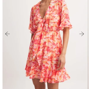 Blommig kortklänning med volang NAKD  - Superfin kortklänning i blommigt mönster med rosa, orange och vita toner. Klänningen har v-ringning, knyt fram och volangdetaljer vid ärmar och fåll. Perfekt flowy passform och elastisk midja för en bekväm känsla.