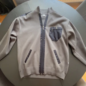 Grå cardigan jacka från Lacoste 3XL - Snygg grå cardigan jacka från Lacoste i storlek 3XL. Jackan har svarta detaljer på axlar, fickor och bröstficka med dragkedja och ikoniska krokodilloggan. Ribbad krage och muddar, hel dragkedja framtill. Perfekt för dig som gillar sportig och clean stil.