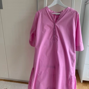 Rosa klänning från Coster Copenhagen - Supersöt rosa klänning från Coster Copenhagen med avslappnad passform, v-ringning och knappar framtill. Klänningen har kort ärm och volangdetalj nertill. Använd 1 gång så nyskick