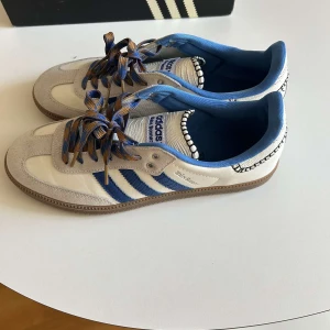 Adidas Wales Bonner Samba sneakers - Unika Adidas Samba Wales Bonner sneakers i vitt och blått med klassiska tre ränder på sidan. Skorna har beige mockadetaljer, blå innerfoder och coola snören i blått och brunt. Snygg kontrastsöm runt hälen och gummisula. Perfekta för dig som gillar limited edition och streetstyle dem är 45 1/5 men passar som 45.