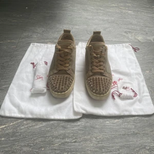 Beige Christian Louboutin sneakers med nitar - Snygga beige sneakers från Christian Louboutin med coola silverfärgade nitar på tån och klassisk röd sula. Skorna har snörning, meshpaneler på sidorna och är tillverkade i mocka för en lyxig känsla. Perfekta för dig som vill sticka ut med exklusiv streetstyle.