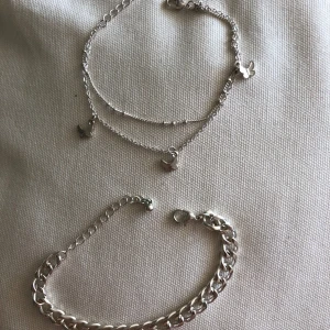 Två silvriga ett fotarmband och en armband med kedjor - Säljer två snygga armband i silverfärg. Det ena är ett två tunnare kedja som sitter ihop med små fjärilsberlocker är ett fotarmband aldrig använd. Det andra är ett klassiskt bredare kedja armband, använd nån gång kanske minns inte. (Inte änta silver någon av dom)