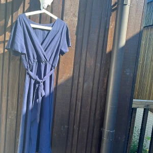 Marinblå omlottklänning med knytband OANVÄND - Snygg marinblå långklänning med omlottliv och v-ringning. Klänningen har korta ärmar och ett knytband i midjan som ger en fin siluett. Perfekt för dig som gillar en enkel men stilren look.