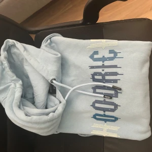 Ljusblå hoodie från Hoodrich XS - Säljer en ljusblå hoodie från Hoodrich i storlek XS. Tröjan har en stor logga framtill i blått och vitt, justerbar huva med snören och en klassisk känguruficka. Perfekt för dig som gillar streetwear och vill ha en chill look.