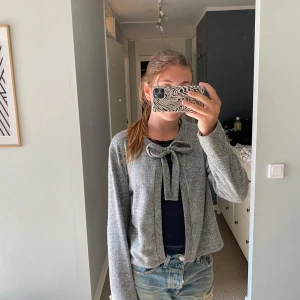 Grå kofta med rosett i ullmix - Superfin grå kofta med knytrosett framtill. Koftan är i mjuk ullmix och har en loose passform med långa ärmar. tröja kommer tyvärr inte till användning längre så hoppas på att någon är intresserad!💛