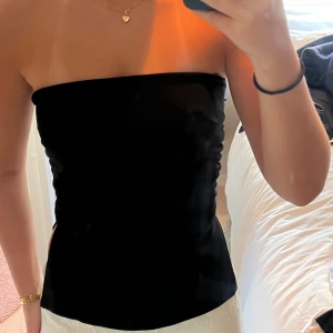 Svart bandeau tubtopp med slits - Säljer en svart bandeau tubtopp med slits på sidan. Toppen är axelbandslös och har en tight passform som sitter snyggt mot kroppen. Perfekt för dig som gillar minimalistisk stil och vill ha en enkel men trendig topp till sommaren.