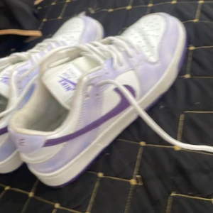 Nike Air Force 1 vit/lila sneakers - Nike Air Force 1 sneakers i vitt och lila med klassisk låg siluett. Skorna har lila swoosh, vita snören och perforerad tå för extra ventilation. Tillverkade i skinn och syntetmaterial, perfekt för dig som vill sticka ut med en fräsch färgkombo.