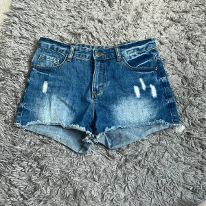 Blå jeansshorts från UNIQ dnm - Snygga blå jeansshorts från UNIQ dnm med slitna detaljer och fransig kant nertill. Klassisk femficksmodell med bälteshällor och knappstängning. 