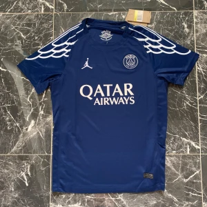 PSG x Jordan blå matchtröja D. Doué - Paris Saint-Germain x Jordan fotbollströja i mörkblått med vita detaljer och vingmönster på axlarna. Tryck med Qatar Airways, PSG-logga och nummer 14 D. Doué på ryggen. Kortärmad och tillverkad i lätt, ventilerande material. Champions League-märke på ena ärmen.