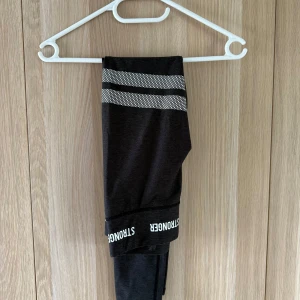 Svarta träningsbyxor från Stronger - Snygga svarta leggings från Stronger med vita loggor i midjan och två vita randiga detaljer på ena benet. Tight passform och stretchigt material som passar perfekt till träning eller chill. Hög midja för extra komfort.