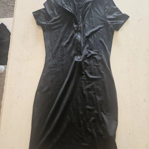 Svart kortklänning med dragkedja från SHEIN - Svart kortklänning från SHEIN i storlek M. Klänningen har korta ärmar, en markerad krage och en dragkedja framtill som ger en cool och sportig vibe. Materialet är mjukt och stretchigt, perfekt för en trendig look.