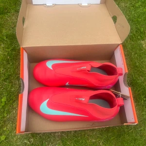 Nike Mercurial fotbollsskor rosa - Snygga Nike Mercurial fotbollsskor i starkt rosa med ljusblå detaljer och vit swoosh. Skorna har täckt snörning och greppiga dobbar för bästa fäste på planen. Lätta och smidiga i syntetmaterial, perfekta för dig som vill sticka ut på matchen. Dem har aldrig används.MVH Ville