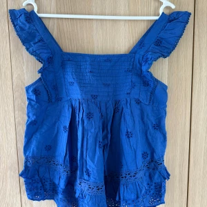 Blå blus med volang från MNG - Supersöt blå blus från MNG med broderade blommor och volangdetaljer på axelbanden och nederkanten. Luftig modell i bomull, perfekt för varma dagar. Blusen har breda axelband och fina hålbroderier som ger en romantisk vibe.