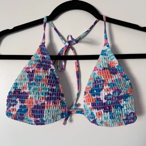 Färgglad bikiniöverdel - Snygg bikiniöverdel med ett färgglatt mönster i lila, blått, rosa och orange. Den har en smockad design och knytband i nacken och ryggen för justerbar passform. Storleken är L men passar bra till mig som har S/M. Perfekt för sommarens stranddagar! 🧡💜 Önskar du veta exakta mått? Tveka inte att höra av dig!