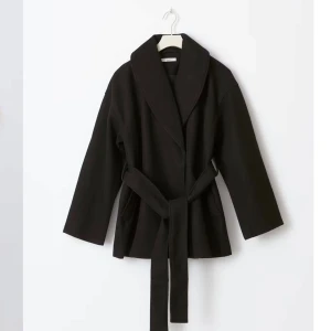 Svart kort kappa med bälte - Snygg svart kappa med bälte och långa ärmar. Perfekt för en stilren look. Den har en enkel design med en knytfunktion i midjan. Har en defekt i fickan (skriv för bild) annars är den i bra skick🩷🩷nypris 700kr