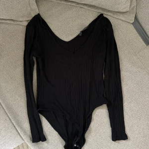 Svart ribbad bodysuit - Säljer en svart ribbad bodysuit med långärmad design och v-ringad hals. Perfekt för lager-på-lager eller att bära som den är. Enkel att matcha med olika outfits.