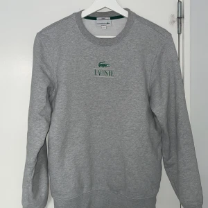 Grå sweatshirt från Lacoste - Säljer en klassisk grå sweatshirt från Lacoste med grönt krokodilemblem och text på bröstet. Tröjan har rund hals och långa ärmar, perfekt för en avslappnad stil. Passar både herr och dam.