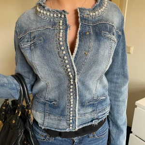 Jeansjacka med pärldetaljer - Snygg jeansjacka med pärlor längs kragen och knappslån. Jackan har en fransig kant och två bröstfickor. Perfekt för att ge din outfit en cool touch. Storlek M men sitter som S-XS