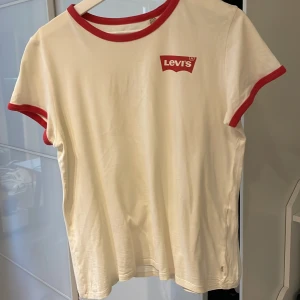 Vit t-shirt från Levi's med röd logga - Säljer en klassisk vit t-shirt från Levi's med röd logga på bröstet. T-shirten har röda kanter vid hals och ärmslut, vilket ger en snygg kontrast. Perfekt för en avslappnad stil.