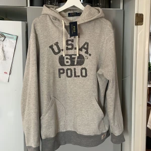 Ralph Lauren Hoddie - Säljer en grå hoodie från Ralph Lauren med trycket 'U.S.A. 67 POLO' på framsidan. Tröjan har en justerbar huva med snören och en stor ficka framtill. Perfekt för en avslappnad stil. Den är helt ny med tags! Ny pris 2,599 mitt pris 799! Inte hugget i sten! Storlek L-M! Kom med bud!😁