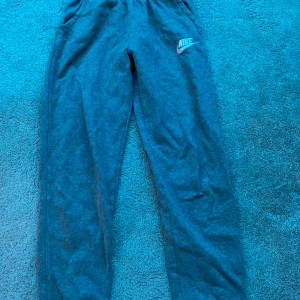 Grå mjukisbyxor från Nike - Säljer ett par grå mjukisbyxor från Nike med elastisk midja och sidofickor. Perfekta för en avslappnad stil eller träning. Nike-loggan är tryckt på ena benet.