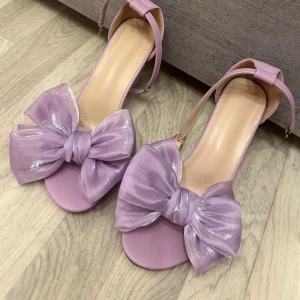 Lila sandaler med rosett - Snygga lila sandaler med en stor glittrig rosett framtill. De har en bred klack och en justerbar rem runt ankeln. Aldrig använda ute utan endast testade hemma.