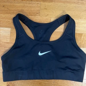 Svart sporttopp från Nike - Säljer en svart sporttopp från Nike med klassisk design och vit logga framtill. Perfekt för träning med sin bekväma passform och elastiska material. Toppen har en racerback för extra stöd och rörelsefrihet.