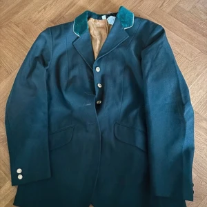 Tävlings kavaj shires - Elegant grön kavaj med kontrasterande grön krage och knappar framtill. Perfekt för en stilren look. Kavajen har långa ärmar och två fickor framtill. 