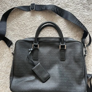 Svart portfölj från Armani  - Snygg svart portfölj från Armani. Den har ett justerbart axelband med logotyp, dubbla handtag och en praktisk dragkedja upptill. Inuti finns flera fack för enkel organisering. Perfekt för att bära med sig det viktigaste med stil. Ny pris 2400 kr.
