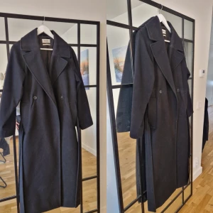 Svart kappa från Weekday - Elegant oversize svart kappa från Weekday med klassisk design. Den har långa ärmar och knappar framtill. Perfekt för kyligare dagar med sin stilrena look.