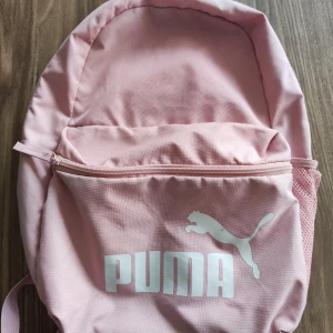Rosa ryggsäck från Puma - Säljer en snygg rosa ryggsäck från Puma. Den har ett stort huvudfack och en praktisk framficka med dragkedja. Axelremmarna är vadderade för extra komfort. Perfekt för skolan eller träningen!