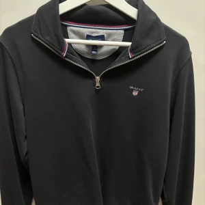 Half zip  GANT - Snygg svart tröja från GANT med dragkedja och broderad logga på bröstet. Tröjan har en stilren design med ribbade detaljer vid kragen och en diskret randig insida. Perfekt för en avslappnad och stilren look.