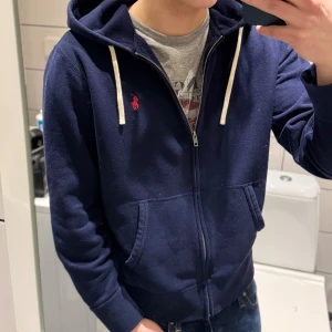 Mörkblå hoodie från Polo Ralph Lauren - Säljer en snygg mörkblå hoodie från Polo Ralph Lauren med dragkedja och det klassiska röda logotypen på bröstet. Tröjan har långa ärmar och en praktisk huva med vita snören. Perfekt för en avslappnad stil. Storlek M! Bara att höra av sig vid frågor och funderingar. Bytesförslag är också intressant!🐎