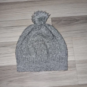 Grå stickad mössa med tofs - Säljer en mysig grå stickad mössa med en fluffig tofs på toppen. Perfekt för kyliga dagar och ger en stilren look. Mössan har en ribbad kant som ger en bra passform. Hemmastickad.