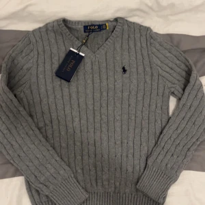Grå stickad tröja från Ralph Lauren - Säljer en stilren grå stickad tröja från Ralph Lauren med kabelstickat mönster och v-ringning. Tröjan har långa ärmar och den klassiska loggan broderad på bröstet. Perfekt för en elegant och avslappnad look.