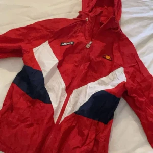 Röd vindjacka från Ellesse - Snygg röd vindjacka från Ellesse med vita och blå detaljer. Jackan har en dragkedja framtill och en huva. Perfekt för en sportig look med Ellesse-logga längs ärmarna.