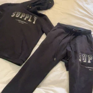 Svart hoodie från Supply & Demand - Säljer en svart hoodie från Supply & Demand med texten 'SUPPLY' tryckt på framsidan. Hoodien har en klassisk design med dragsko i huvan och ribbade muddar vid ärmslut och nederkant. Perfekt för en avslappnad stil.