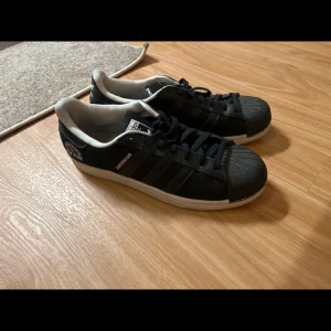 Svarta Adidas Superstar sneakers - Snygga svarta Adidas Superstar sneakers med vit sula och klassiska tre ränder på sidan. Skorna har texten 'THE KAISER OF NEW YORK' på hälen och Adidas-logga baktill. Perfekta för en stilren look.