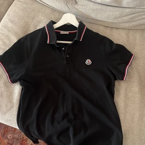 Svart pikétröja från Moncler - Snygg svart pikétröja från Moncler med klassisk krage och knappar. Tröjan har Monclers logotyp på bröstet och röd-vita detaljer på krage och ärmslut. Perfekt för en stilren look.