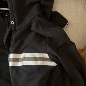 Svart hoodie från Moncler - Säljer en stilren svart jacka från Moncler med huva och dragkedja. Jackan har två vita ränder på ärmen och ribbade muddar. Perfekt för en cool och avslappnad look.