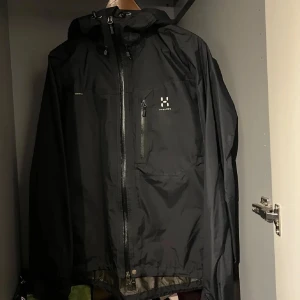 Svart regnjacka från Haglöfs - Säljer en svart regnjacka från Haglöfs med dragkedja och justerbar huva. Jackan har en bröstficka med dragkedja och är perfekt för regniga dagar. Den är både funktionell och stilren.