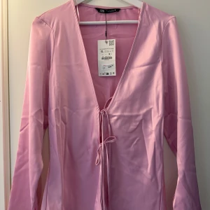 Rosa långärmad topp från Zara - Säljer en stilren rosa långärmad topp från Zara. Toppen har en knytning framtill och är tillverkad i ett glansigt material som ger en elegant look. Aldrig använd med prislappen kvar. Liten i storlek, passar M/L. 