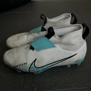 Nike Mercurial fotbollsskor - Säljer ett par vita Nike Mercurial fotbollsskor med turkosa detaljer och svart Nike-logga. Skorna har en modern design med snörfri passform och en elastisk rem över vristen för extra stöd. Perfekta för snabba rörelser på planen.