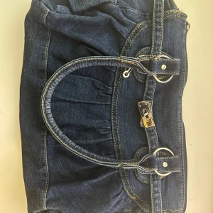 Blå jeansväska från Kappahl - Snygg blå jeansväska från Kappahl med dubbla handtag och dekorativa spännen. Väskan har en praktisk dragkedja för säker förvaring. Perfekt för en casual look och rymmer allt du behöver för dagen. Tyvärr är låset borta men kan lätt bytas ut 
