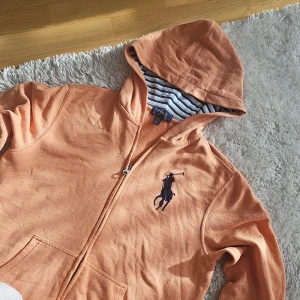 Orange huvtröja från Ralph Lauren - Säljer en snygg orange huvtröja från Ralph Lauren med dragkedja och stor broderad logga på bröstet. Tröjan har en randig insida i huvan och är långärmad. Perfekt för en avslappnad stil. Storlek barn 14-16 år men passar perfekt för vuxna storlek S-M.