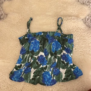 Blommig linnetopp med blå rosor - Säljer en söt linnetopp med ett blommigt mönster av stora blå rosor och gröna blad. Toppen har smala axelband och en luftig passform, perfekt för varma dagar.