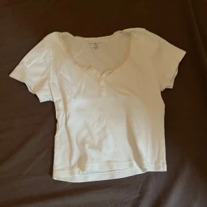 Söt topp med spetsdetalj - Säljer en söt vit topp från brandy melville med korta ärmar och spetsdetalj vid halsringningen. Toppen har knappar framtill och är i en mjuk bomullskvalitet. Perfekt för en avslappnad stil.