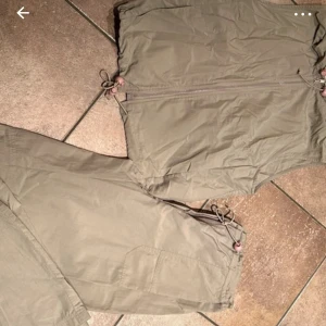Beige fritidssett - Säljer en praktisk beige väst med dragkedja framtill. Västen har justerbara snören i nederkant för en anpassad passform. Även matchande långa byxor till. Perfekt för utomhusaktiviteter.