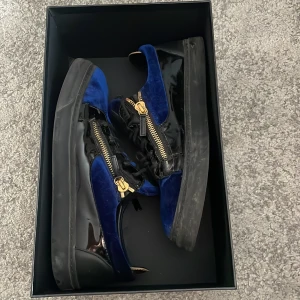 Svart och blå sneakers från Giuseppe Zanotti - Snygga sneakers från Giuseppe Zanotti i svart och blått med guldfärgade dragkedjor. Skorna har en kombination av glansigt material och sammet, vilket ger en lyxig känsla. Perfekta för den som vill sticka ut med stil. Kan tvätta dom innan köp