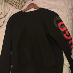 Svart tröja från Hugo - Snygg svart tröja från Hugo med röd logga på ärmen och bröstet. Perfekt för en stilren look. Tröjan har långa ärmar och en bekväm passform.